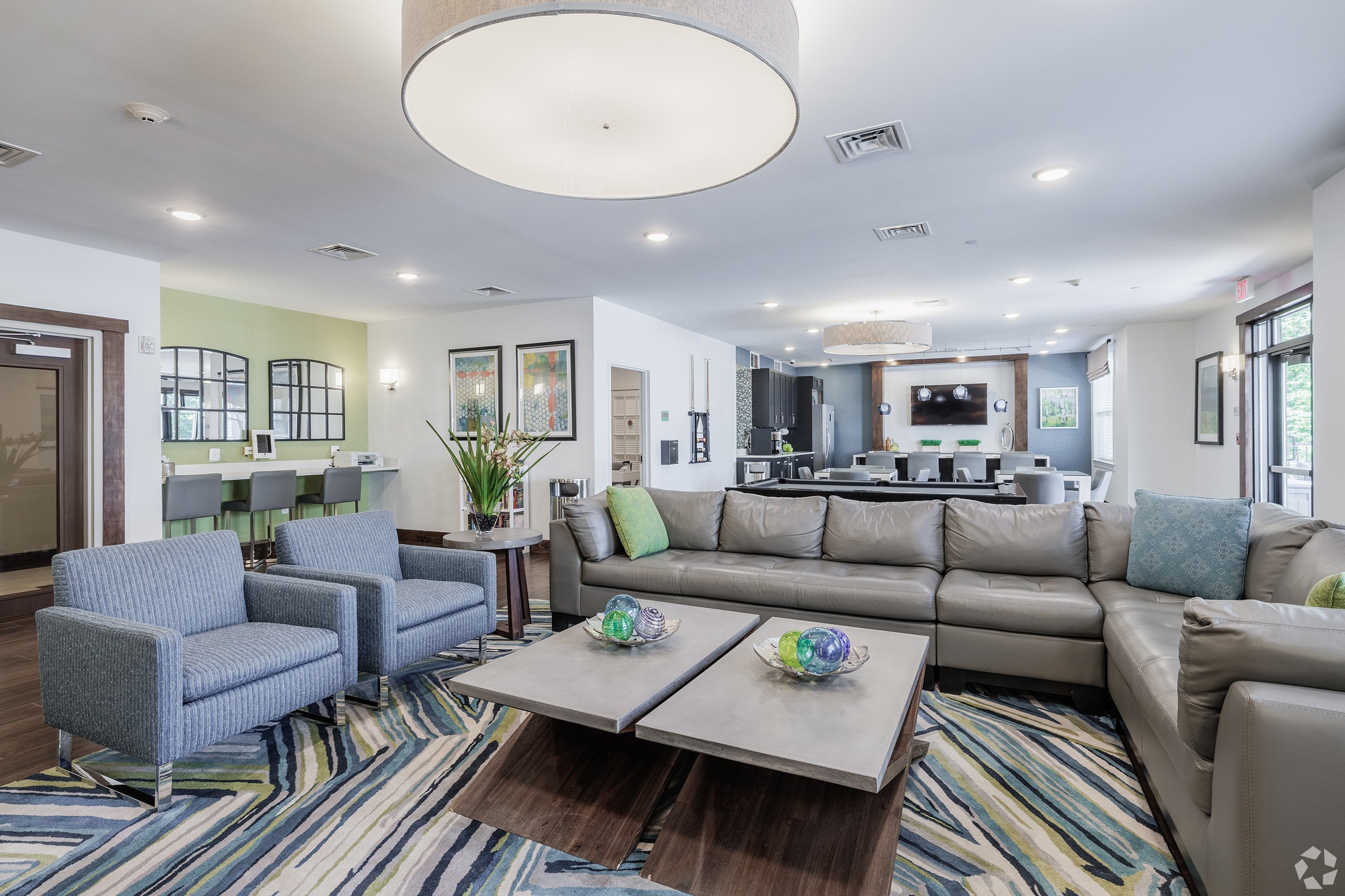 Residents' lounge with three seating areas, leather sectional sofa, accent chairs, dining bar, large pendant light, mounted TV, and woodland-patterned rug
